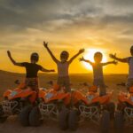 Group of friends enjoying a quad biking tour at sunset in the Agafay Desert near Marrakech, riding orange ATVs with arms raised in celebration against a golden sky.