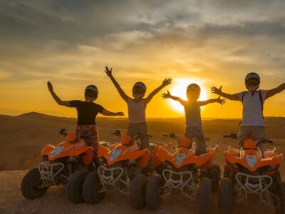 Group of friends enjoying a quad biking tour at sunset in the Agafay Desert near Marrakech, riding orange ATVs with arms raised in celebration against a golden sky.